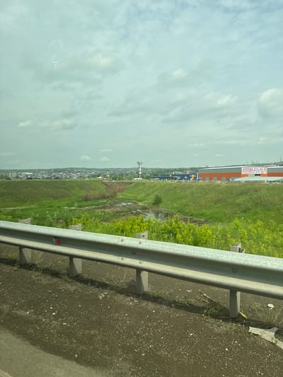 An outdoor, eye-level, wide shot captures a guardrail in the foreground with a rural-urban landscape in the background under an overcast sky. The guardrail, made of metal, is mounted on concrete posts, and there is a red reflector attached to the left side of the rail. The ground beneath the guardrail is dark and rocky, with sparse green vegetation growing through it.
Beyond the guardrail, a green, grassy hillside slopes down towards a small, muddy area with some scattered vegetation. Further in the background, a city skyline is visible with various buildings and a tall tower with "METRO" written on it. There is also a large building with orange and grey panels and a sign that reads "АРЕНДА 214-44-44" and "ПРОЕКТИРОВАНИЕ И СТРОИТЕЛЬСТВО 2-71-22-11". The overall scene suggests a location on the outskirts of Солонцы, Russia, where a rural area meets a developed commercial or industrial zone. No people are visible in the image.