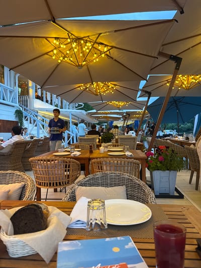 The image depicts an outdoor dining area at a restaurant in Zhukovka, Russia, likely during twilight. Numerous large beige umbrellas, adorned with strings of warm yellow lights, extend over several wooden tables set for dining. The tables are occupied by patrons and are furnished with wicker chairs, plates, glasses, and napkins. In the mid-ground, a waiter in a blue shirt and grey pants carries a tray of plates. A white building with a staircase and white railing is visible in the background, and a body of water can be seen further back, suggesting a waterfront location. The overall ambiance is relaxed and inviting, typical of an evening meal at a resort town.