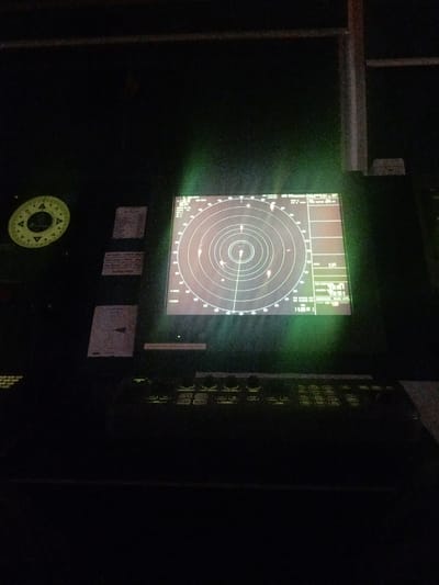 The image depicts a dimly lit bridge of a ship, likely at night or in low-light conditions, as indicated by the dark surroundings and the illuminated screens. The primary focus is a radar display, showing concentric circles representing ranges and several blips indicating other vessels or objects. The radar screen has a greenish hue, typical of older radar systems.
To the left of the radar, there's a circular instrument with yellow markings, possibly a gyrocompass repeater or a bearing indicator. Below this, several rectangular placards with text are attached to the console; however, the text is too blurry to be legible.
In front of the radar screen is a keyboard or control panel with numerous buttons, many of which are illuminated with a yellow-green light. Some of the visible labels on the control panel include "POWER," "OFF," "ON," and others that are indistinct. The overall impression is one of a functional control station within a vessel, where navigation and monitoring are taking place. The scene suggests a sense of quiet operation, with the glow of the instruments being the main source of light. There are no people visible in the frame. The environment appears to be an interior, enclosed space.
