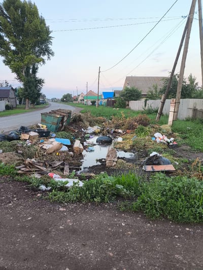 The image depicts an outdoor scene in Saraktash, Russia, characterized by a significant accumulation of garbage and debris. Two large, green dumpsters are positioned near a dirt road, overflowing with trash. Scattered around the dumpsters is a large pile of various waste items, including plastic bags, cardboard boxes, dry grass, and what appears to be a pool of stagnant water. The surrounding environment shows a rural or suburban setting with houses and trees visible in the background. Power lines stretch across the sky. The overall impression is one of neglect and unsanitary conditions.