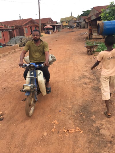 This image depicts a vibrant street scene in Isieke, Nigeria, under what appears to be an overcast daytime sky. The setting is an unpaved road characterized by reddish-brown dirt and scattered small stones, with buildings lining its edge.
In the foreground, a man is seated on a blue and white motorcycle, likely a moped or small utilitarian model, which shows signs of wear. He is dressed in a khaki-colored short-sleeved shirt and dark trousers or shorts, wearing sandals. He holds the handlebars and looks directly towards the viewer, appearing calm.
Slightly behind and to the right of the man on the motorcycle, a younger person or child stands on the dirt road, facing away from the camera. This individual is actively carrying a large, blue gas cylinder, which rests on their left shoulder. They wear a light-colored short-sleeved t-shirt with the text "S.F.N.P.O. ISIEKE" visible on the back, and light-colored checkered shorts.
The background features a row of buildings, suggesting a local community or commercial street. These structures predominantly have reddish-brown corrugated iron roofs, with walls of various colors, some painted orange or red. Sparse vegetation, including trees, and a utility pole are also visible, further defining the rural or semi-urban context. The scene captures a moment of everyday life, highlighting modes of transport and daily tasks in the area.