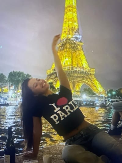 A low-angle, medium shot captures a young woman with long dark hair, wearing a black t-shirt that reads "I ❤️ PARIS" and jeans, reclining on a stone surface next to a body of water. Her right arm is raised, and she is smiling upwards. In the background, the Eiffel Tower is illuminated at night, its golden lights reflecting on the water. Several boats are visible on the water, and trees and other structures line the distant shore. A dark bottle, possibly wine, is on the stone in the foreground to the left, along with two white paper cups. A person's legs and feet, wearing dark shoes and white socks, are visible on the right side of the frame. The scene appears to be a nighttime setting with artificial lighting from the Eiffel Tower and other sources. The image does not depict the city of Kołobrzeg, Poland; the Eiffel Tower is located in Paris, France.