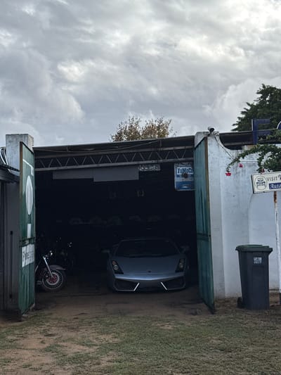 The image shows the interior of a garage or storage area in Kraaifontein, South Africa. Inside, there's a grey Lamborghini and a motorcycle. A green garbage bin is visible near the entrance. Signs are mounted on the structure. The sky is overcast with heavy clouds. The surrounding ground appears to be dirt and sparse vegetation.
