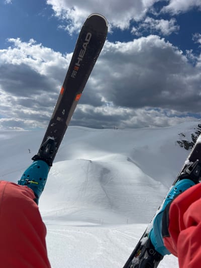 The image displays a first-person perspective from an individual wearing skis, situated in the city of Katun Bačko brdo, Montenegro. Two dark gray "PRO HEAD" brand skis, exhibiting orange accents and minor snow/ice accumulation, extend into the lower frame. Each ski is secured to a light blue ski boot, which covers the foot of a person wearing orange ski pants. The background presents an expansive snow-covered mountainous terrain, featuring discernible ski tracks and groomed sections. At least two distant ski lift towers are visible. Darker, rocky patches are present on the far right. The sky above shows a mix of large, dark gray cumulus clouds and patches of clear blue. The scene is illuminated by daylight.