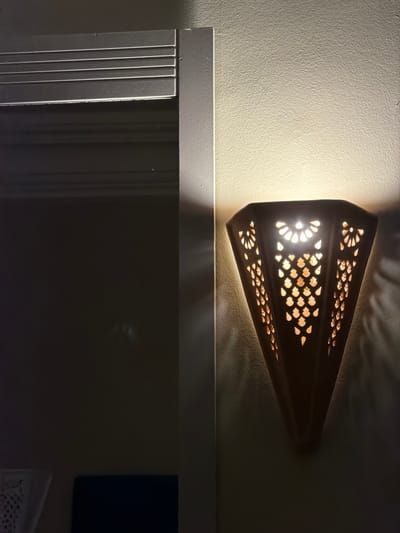 The image shows a wall-mounted lamp with an intricate cutout design, casting a warm, patterned light onto the textured cream-colored wall. The lamp itself is a dark, conical shape, wider at the top and tapering to a point. The light source within illuminates the cutouts, which resemble stylized floral or teardrop shapes, creating a dappled effect on the wall. To the left of the lamp, a portion of a dark, recessed area is visible, featuring horizontal ridged detailing on its surface, suggesting a doorway or cabinet. The overall scene is dimly lit, with the lamp being the primary light source, creating a contrast between illuminated and shadowed areas. There are no people or animals visible in the image. The setting appears to be an indoor space, possibly a hallway or a room in a home. Without further context, the specific location within the city of Sousse, Tunisia cannot be determined from the visual information alone.