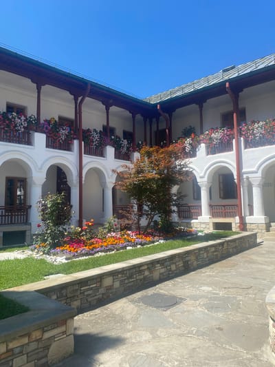 The image shows an interior courtyard of a white, multi-story building with arched walkways and a covered balcony lined with potted flowers. In the center of the courtyard is a small tree with reddish-brown leaves and a flowerbed filled with a variety of colorful blooms. The ground of the courtyard is paved with irregular stone slabs, and a low stone wall borders the flowerbed and a grassy area. The sky above is clear and blue. The building appears to be part of a monastery or a similar historical structure, likely the Agapia Monastery in Agapia, Romania, given the provided context. No people are visible in the image.