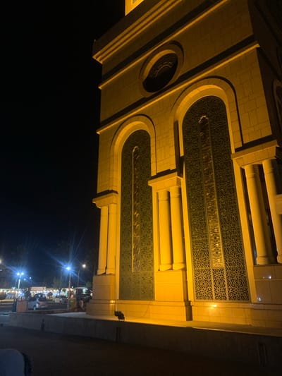 The image, captured at night in Samarinda, Indonesia, showcases the ornate facade of a building, likely a mosque or cultural edifice, bathed in warm, artificial light. The structure features light-toned, possibly tiled, construction elements that appear golden. Key architectural details include prominent rectangular frames, gracefully rounded archways, and large panels filled with intricate dark geometric patterns characteristic of Islamic design. Integrated into these patterned sections are horizontal strips of reflective material, within which decorative elements and faint, unreadable Arabic calligraphy are hinted at. Substantial cylindrical pillars or decorative columns are also visible, adding to the building's grandeur.

To the right and below the illuminated building, a vibrant street scene unfolds. Multiple bright streetlights cast starburst flares and illuminate the area, revealing parked vehicles and a few indistinct figures, suggesting an urban environment with ongoing activity, possibly market stalls or shops. The sky overhead is dark, emphasizing the striking contrast between the richly lit architecture and the nocturnal surroundings. The image conveys a sense of serene beauty and cultural depth within a bustling urban nightscape.