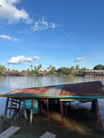The image presents a riverside scene in Khorn Neua, Laos, under a bright blue sky with scattered white cumulus clouds. In the foreground, a partially submerged wooden structure is visible, featuring a corrugated metal roof composed of alternating sections of rusty red and galvanized silver. The structure's posts, painted in red, green, and blue, descend into the murky brown river water, with parts of the lower framework fully submerged. Two weathered wooden planks rest on the muddy bank in the extreme lower-left corner. The wide, brown river occupies the midground, showing minor ripples on its surface. The far bank displays a settlement with numerous traditional buildings and stilt houses nestled among abundant palm trees and dense green foliage. Several tall red and white communication towers punctuate the skyline above the village. No individuals or direct human activities are depicted in the scene.