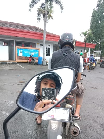 A man in a motorcycle helmet takes a selfie in the motorcycle's side mirror. The reflection shows his face and phone. The main view reveals the back of another man, also in a helmet and vest, on the same motorcycle. They're stationary in a group of motorcycles. In the background, a low building with a red roof and a palm tree are visible. The scene appears to be outdoors on a cloudy day.