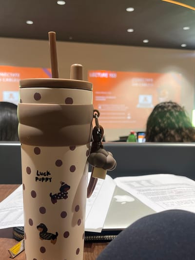 The scene depicts an indoor lecture or classroom setting located in Fraccionamiento Los Heroes Queretaro, Mexico.
In the immediate foreground, a beige water bottle with brown polka dots and a brown silicone grip band occupies the central frame. The bottle features the text "Luck Puppy" and illustrations of two dachshunds. It has a brown lid with a visible straw and a small stopper. A brown mushroom-shaped charm hangs from a strap attached to the bottle. The bottle rests on a dark wooden surface. To the left, a portion of a black zippered pouch and a small yellow object are visible. To the right, an open book or notebook displaying text is present, with the faint reflection of an Apple logo visible beneath it, likely from a laptop or tablet. A segment of dark blue fabric, presumably clothing, is visible in the bottom right corner.
In the midground, blurred, the backs of two individuals are visible. The individual on the left has dark hair, while the individual on the right displays curly, green-tinted hair. Both appear to be seated in front of desks or partitions.
The background features a large, blurred projection screen displaying an orange background with white text. Partially legible text includes "LECTURE 103" and "CONECTATE UN CABLE," accompanied by a graphic of a laptop. Recessed ceiling lights are visible above the screen. The overall lighting is artificial.