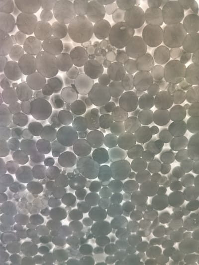 The image is a close-up, abstract shot of a white, granular surface that appears to be styrofoam. The granular texture is composed of numerous small, roughly spherical beads that are densely packed together. The lighting is from behind the surface, causing the beads to be backlit and appear as a collection of circles with varying degrees of opacity and shadow. Some beads are well-defined circles, while others are softer or show internal texture. The overall color palette is monochromatic, with shades of grey and white, and a subtle gradient that shifts from a cooler, bluish-grey on the left to a warmer, yellowish-grey on the right. The image does not contain any discernible people, objects, or actions that would suggest a specific location in the city of Parabel', Russia, or any other identifiable urban environment. It is purely an abstract textural image.