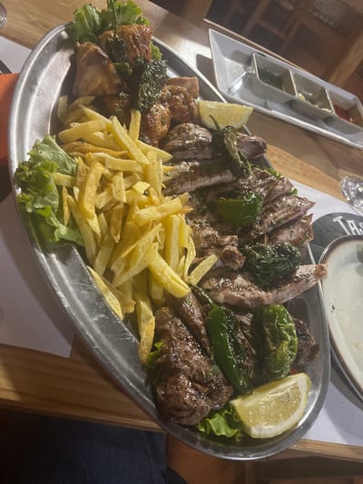 A close-up, overhead shot captures a large, metallic platter filled with grilled meats, french fries, and salad. The platter is oval-shaped and appears to be made of stainless steel. The food is arranged in a somewhat chaotic, yet appetizing, manner.

Dominating the left side of the platter are generous portions of golden-brown french fries, some still appearing crisp. To the left and back of the fries are fresh, green lettuce leaves, providing a crisp contrast to the fried potatoes.

The right and central areas of the platter are covered with various grilled meat cuts. These meats are sliced and appear to be a mix of dark and light brown, with visible grill marks. Scattered amongst the meat are several bright green chili peppers, adding a pop of color and suggesting a spicy element. A slice of lemon is visible on the bottom right of the platter, offering a zesty garnish.

In the background, a white rectangular tray with three compartments holds various dipping sauces, possibly a green herb sauce, a lighter colored sauce, and a red sauce. The tray is positioned behind the platter of food. The setting appears to be a restaurant, with wooden tables and chairs visible in the blurred background. The lighting suggests it might be evening.

The location of Las Dehesas, Spain, is not explicitly depicted in the image, but the presence of a meal being served implies a dining establishment within that locale. No individuals are visible in the image. The scene is focused entirely on the prepared food on the platter.