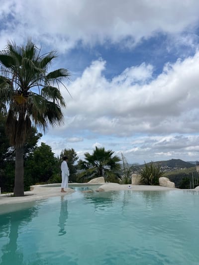 The image depicts a serene outdoor scene, likely in Can Güell, Spain. In the foreground, a clear, turquoise infinity pool dominates the lower half of the frame, reflecting the sky and its surroundings. A person, seen from the back, stands on the pool's edge, wearing a white robe. To the left of the person, a tall palm tree with dark fronds stands prominently. The background reveals a lush, green hillside dotted with houses, suggesting a residential area nestled within nature. The sky above is a dramatic canvas of blue, interspersed with large, fluffy white clouds, hinting at a partly cloudy day. The overall atmosphere is one of tranquility and natural beauty.