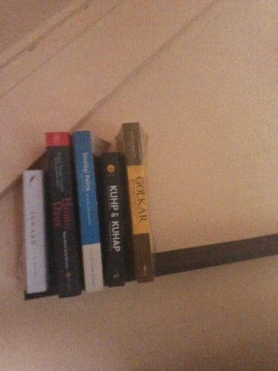 A slightly grainy, static image depicts five books standing upright on a dark, narrow shelf against a light, textured wall in Dogsenang Gorphoog, Bhutan. From left to right, the book spines are: a white book titled 'JAWAD', a black book with a red top edge titled 'Homo Deus', a blue book with white trim titled 'Strategi Politik', a dark book displaying a yellow circular emblem and 'KUHP & KUHAP', and a dark book with a yellow section bearing 'GOLKAR'. The shelf casts a subtle shadow to its right. No individuals are present, and no actions or interactions are depicted within the frame.