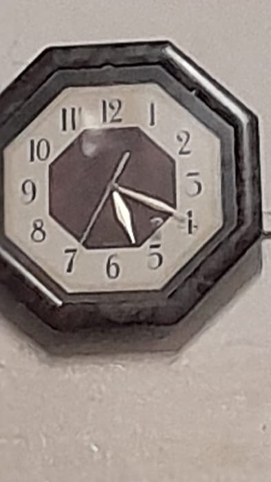 An octagonal analog clock with black hands and a white face is mounted on a textured light gray wall. The hour hand points to the 2, the minute hand points to the 4, and the second hand points to the 4. The clock is located in the city of Merlo, Argentina, at latitude -34.685 and longitude -58.768. The clock appears to be in working order.