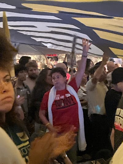The image depicts a dense outdoor gathering of people in an urban environment in West Jerusalem, Israel, under artificial illumination. An overhead canopy features a blue and white striped pattern with yellow accents. In the mid-ground, a person with short dark hair, wearing a red t-shirt inscribed with partially visible text "FIGHTING FOR OUR HOME. TOGETHER WE STAND" and a white scarf, raises their left arm with an open hand. A white badge is visible on their shirt. To their left, a person with dark curly hair and glasses is partially visible in the foreground. In the background, a man holds a black and white sign aloft, displaying the text "NOUS ALLI." Numerous other individuals are visible throughout the frame, forming a crowded scene, with several arms raised among them.