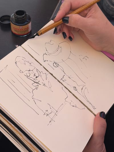 A close-up overhead shot shows a person's hands and an open sketchbook with pen-and-ink drawings on a dark surface. The sketchbook is lying flat, open to a two-page spread. The left page contains a sketch of a figure, possibly a person, with rough lines defining the head and body. The right page features a sketch of what appears to be a hat and more abstract linework. A small, dark glass bottle labeled "INDIAN INK" is visible in the upper left corner, next to the sketchbook. A wooden dip pen, dripping with ink, is held by the person's right hand, poised over the sketchbook. The person's fingernails are painted with dark, glossy nail polish. The background is a dark, textured surface, possibly a table. The image does not contain any information or context to suggest a location in the city of Vernon, France.