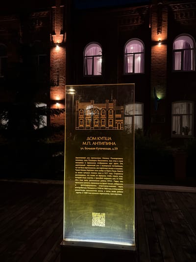 The image captures a brightly illuminated historical information sign at night, set against the dark facade of a brick building in Nikolo-Beryozovka, Russia. The sign, made of a translucent material with an internal light source, casts a yellow-green glow, highlighting its Cyrillic text and a line-art illustration of a two-story house.
The building behind the sign is constructed of dark brick, with several windows featuring white frames and light-colored curtains or blinds, revealing interior lighting. Wall-mounted lamps on the facade cast warm light onto the brickwork. The overall scene is dark, characteristic of nighttime, with artificial lights providing the main illumination.
The visible text on the sign, "ДОМ КУПЦА М.П. АНТИПИНА ул. Большая Купеческая, д.59" (House of Merchant M.P. Antipina, Bolshaya Kupecheskaya Street, 59), details the history of the property. It states that the two-story brick house belonged to Mikhail Polikarpovich Antipina, built as a mirror image of his father's house. The property included outbuildings, a granary, two brick warehouses, a cellar, and a barn by the Kama River. Merchants left the village in 1917, and Antipina reportedly returned later to retrieve a treasure, which he gave to authorities requesting the house's preservation. From the 1920s to 1940s, it housed the district committee of the Communist Party (райком КПСС), where rallies and other public events took place. Between 1958 and 1968, it briefly served as an eight-year school before reverting to the party committee. The text concludes that relocation began in 1980, and the village became deserted. A QR code is present in the lower right corner of the sign. The sign itself shows some visible scratches on its surface. No people or activities are explicitly depicted; the scene is static, focusing on the historical monument.