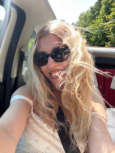 In the city of Schoenfels, Luxembourg, a woman with blonde hair and sunglasses is in a car. She is wearing a white lace top and a black seatbelt. Her hair is blowing in the wind, and she is smiling. The car appears to be a convertible, with the top down, as evidenced by the trees and sky visible in the background. The interior of the car is visible, with red seats in the back. The scene is brightly lit by the sun, with some lens flare present in the upper left corner of the image.