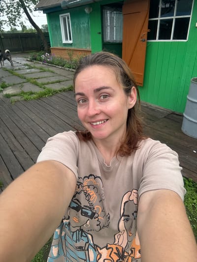 A medium-shot selfie shows a young woman with brown hair and light skin, wearing a beige t-shirt with a graphic print of two women. She is smiling at the camera. The background is a wooden deck, with a green building with orange shutters and windows behind her. There is a dog visible in the distance to the left, and a gray barrel on the right. The scene appears to be a residential yard, likely in a rural or suburban setting. The image's metadata indicates it was taken in Gonja, Russia.