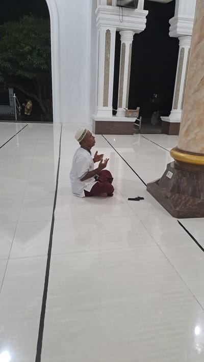 A middle-aged man, appearing to be of South Asian descent, is captured in a moment of prayer, seated cross-legged on a polished white tiled floor. He wears a white skullcap and a short-sleeved white tunic over maroon trousers. His hands are raised with palms facing upwards, indicative of supplication. The setting appears to be an open-air atrium or covered walkway, possibly part of a mosque, given the architectural elements visible. White decorative pillars with beige accents frame an archway in the background, beyond which darkness suggests it is nighttime. To the right, a large, fluted column with a golden capital stands prominently. The floor is sectioned by thin black lines. A small, dark object, possibly a phone or other electronic device, lies on the floor near the man's right side. In the far background, through an arched opening, a person in a light-colored garment is visible standing near a tree. The overall atmosphere is one of quiet devotion within a structured, possibly religious, architectural space in Lhokawe Teungoh, Indonesia.