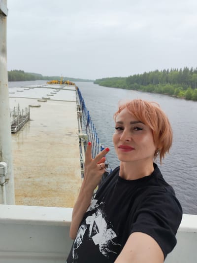 The media file shows a woman taking a selfie on a boat. She has short, reddish-blonde hair, dark eyeliner, and red lipstick. She is wearing a black t-shirt with a white graphic design. Her left hand is raised, displaying a peace sign with red nail polish. She is also wearing a ring on her left ring finger.
The background shows a wide, calm river with tree-lined banks on either side, suggesting a natural, possibly rural or forested, environment. The sky is overcast and grey, indicating a cloudy or possibly rainy day. The boat appears to be a large vessel, possibly a barge or cargo ship, with a wide, flat deck. Part of the deck is visible in the background, with some structures and railings. The overall impression is one of a serene, albeit slightly dreary, day on the water. The location is identified as Annensky Most, Russia.