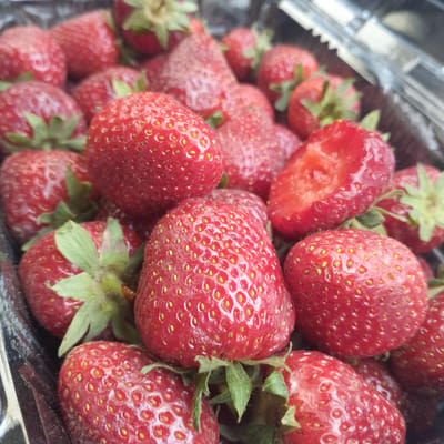 A close-up, high-angle shot captures a cluster of fresh, ripe red strawberries in a clear plastic punnet. The strawberries fill most of the frame, showcasing their vibrant color and textured surfaces. Many of the berries still have their green calyxes and short stems attached, indicating freshness. Distinct yellow achenes (seeds) are clearly visible across the bright red skin of several strawberries.
In the mid-right of the image, one strawberry appears to be bitten or cut open, revealing its uniformly red interior flesh. The plastic container has ribbed edges and a slightly glossy appearance, with reflections suggesting ambient light. The overall impression is one of abundance and ripeness. The lighting is natural and bright, suggesting it is daytime. No people or visible text are present. The scene is set in Обшорон, Tajikistan.