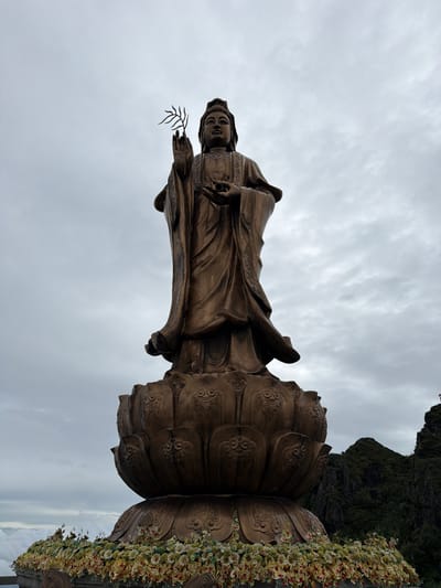 This media file showcases a large, bronze statue of a female figure, likely a deity, with intricate detailing on her robes and face. She holds a delicate sprig in her right hand. The statue is positioned atop a colossal lotus flower base, also bronze, adorned with decorative patterns. Artificial flowers, in shades of yellow, white, and red, cascade down the side of the lotus base, adding a vibrant contrast.

In the background, lush green mountains rise dramatically against a cloudy, overcast sky. The scene appears to be set outdoors in a mountainous region. The cloudy weather suggests it could be daytime, but the light is diffused and somewhat somber.

The image is oriented with the statue lying on its side, making it appear as if the viewer is looking at it from a low angle and sideways. The overall impression is one of grandeur and spiritual significance, set against a natural and somewhat dramatic landscape. No text is visible in the image.

The location is identified as Bản Bo, Vietnam, which is known for its mountainous terrain and cultural sites. The statue's style suggests a Buddhist or East Asian religious context.