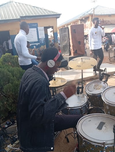 The image captures an outdoor musical performance in Jebbu Miango, Nigeria. In the immediate foreground, a male drummer, wearing a dark denim jacket over a light patterned shirt and dark trousers, is seated, actively playing a drum kit. He wears red-and-white over-ear headphones with an "S" logo and glasses. His right hand, holding a drumstick, is blurred with motion above a snare drum. The drum kit includes at least three tom-toms, a snare drum, and two cymbals, one visibly branded "Sabian sbr." Audio cables and a power strip are on the ground near the drummer's left.
To the left in the mid-ground, a male guitarist or bassist, clad in a white long-sleeve turtleneck and blue jeans, is seated and playing a stringed instrument. To the right in the mid-ground, a male vocalist wearing a white long-sleeve shirt with a logo on the chest and dark trousers stands, holding a microphone to his mouth. Two large, dark rectangular PA speakers, one with a brown fabric covering, are positioned behind the performers.
The background features two buildings with corrugated metal roofs. The structure on the left, painted light brown and blue, displays a prominent white sign with blue text, advertising "First Bank" and "you can more with access" services, including options for e-payment, money withdrawal, transfer, bill payment, and airtime. The building on the right is a multi-story structure with visible wooden balcony railings. The far background to the right reveals a bustling market or street scene with numerous indistinct figures and parked motorcycles. A green shrub is present to the left of the drummer. The ground surface appears unpaved, consistent with an outdoor public area. The scene is brightly lit by natural daylight.