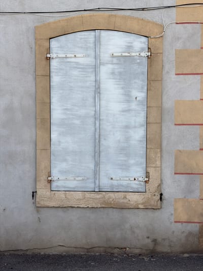 The image shows a closed window with light blue shutters on the exterior of a building in Mercurol-Veaunes, France. The window is framed by a light brown stone arch and sill. The shutters are made of vertical wooden planks and are held closed by two white metal hinges on each side. The wall to the left of the window is gray and textured. To the right of the window, the wall is also gray but features a pattern of alternating beige and red rectangular blocks. Electrical wires are visible running along the top of the wall and connecting to a junction box. The ground in front of the building appears to be asphalt. There are no people visible in the image.