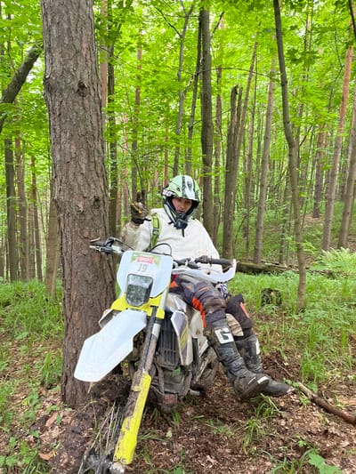 A man wearing a helmet, motocross boots, gloves, and motocross gear is lying on the ground next to a dirt bike. The dirt bike appears to have crashed or fallen over. The scene is a wooded area with trees and greenery. The location is in Gruzinskii, Russia.