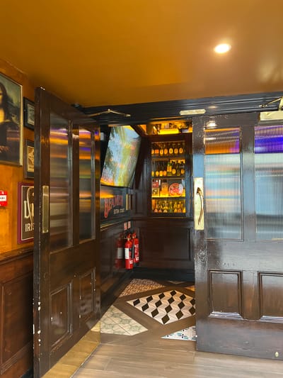 The image depicts an open wooden door, revealing a display of bottles and a sign with the text "BEAMISH EXTRA STOUT" in bold red letters against a black background. Two red fire extinguishers are visible on top of the door. The scene appears to be an interior, possibly a pub or bar, with warm lighting and decorative tiling on the floor. The overall impression is one of an inviting establishment, likely in Donegal, Ireland, given the context. The time of day is not explicitly indicated, but the interior lighting suggests it could be evening or a dimly lit interior. There are no people visible in the frame.