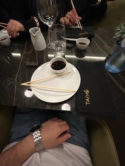 The image captures a dining table setup in a restaurant, likely an upscale sushi establishment given the presence of items such as chopsticks, soy sauce bowls, and a black table mat with the name "TAIYŌ" embroidered on it. The table has a black marble or faux marble surface. Two individuals are partially visible in the frame. One on the left, wearing a dark-colored outfit, is holding chopsticks, suggesting they are in the process of eating. A water bottle and glassware are nearby. On the right, another person's arm and part of their torso can be seen; they are wearing a white shirt and blue jeans, and a silver wristwatch is on their wrist. The location is unspecified and unconfirmable within the city of Chaves, Portugal. The focus of the image is on the dining experience, showcasing the table setting and the people interacting with it.