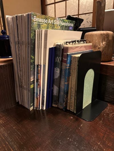 A stack of books and magazines are leaning against a dark wooden wall in a room in Naoshima, Japan (34.458, 133.995). There are at least eleven books and magazines in total. The books on top are in Japanese. Some are hardback and some are paperbacks. The magazines are all identical. The top magazine says "Benesse Art Site Naoshima" and "Wonderful Setouchi." The books are being held in place by a black bookend. There is also a wooden object in the top left of the image. The wooden wall has a light brown wood grain pattern. The background of the image is out of focus.