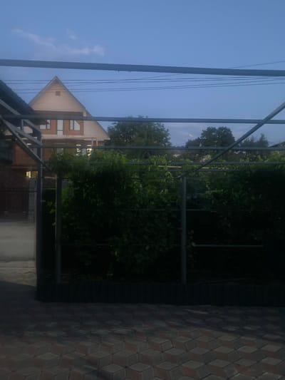 The image shows an outdoor scene with a house, trees, and a paved area. The house has a light-colored facade and a triangular roof with windows. It appears to be a multi-story building. Behind the house, there are dense green trees.
In the foreground, there's a metal structure, possibly a pergola or a part of a building's frame, with some thin wires running vertically. The paved area to the right is made of hexagonal tiles in varying shades of brown and gray, creating a geometric pattern. The overall lighting suggests it might be dusk or dawn, with the sky being a soft blue. The weather appears calm and clear.
There are no people visible in the image, and no text is present. The setting seems to be a residential area in Besagash, Kazakhstan. The mood of the image is quiet and serene, with the natural elements of trees and sky contrasting with the man-made structures of the house and pavement. The composition is dominated by vertical lines from the metal structure and wires, and the angularity of the house and paving.