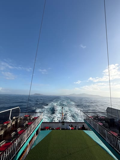 The image shows an ocean view from the deck of a boat, likely taken during a trip from Corralejo, Spain, judging by the distant island. The blue sky with scattered clouds occupies a significant portion of the frame. The sea is rough, with white-capped waves. Part of the boat’s deck is visible on both sides of the image, with rows of empty chairs. There are people in the midground.
