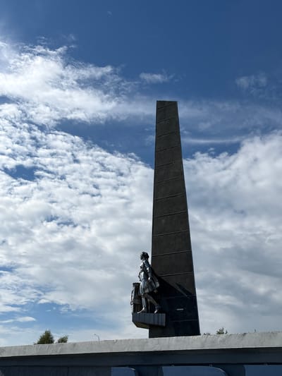 A monument with a tall, dark, obelisk-shaped structure stands against a bright blue sky with scattered white clouds. A silver sculpture of a woman walking is positioned at the base of the obelisk. The monument is situated on a concrete platform, with more concrete structures visible in the foreground. Trees are visible in the background. This is the monument "Per Aspera Ad Astra" in Kirovo-Chepetsk, Russia.
