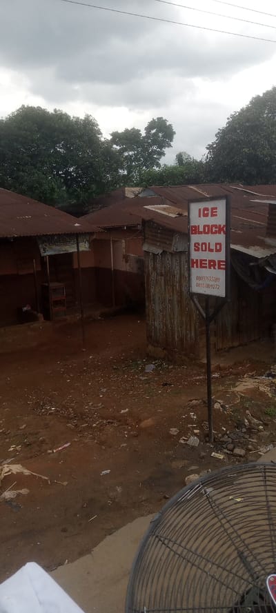 This image displays a semi-urban environment in Ezinifite, Nigeria, under an overcast sky. The foreground consists of reddish-brown dirt ground, uneven and littered with small debris and sparse vegetation. In the lower right corner, a portion of a black metal fan guard is visible, and in the lower left, a white object, possibly fabric.
A prominent black-framed sign stands in the mid-ground on the right, featuring a white face with "ICE BLOCK SOLD HERE" printed in red capital letters. Below this text are two phone numbers: "08037475598" and "08060109272". This sign is positioned in front of a small, weathered structure constructed from heavily rusted corrugated metal sheets.
Behind and to the left of the sign are multiple single-story buildings, primarily featuring aged and rusted corrugated metal roofs. The walls of these structures are painted in earthy tones, predominantly brown and reddish-orange. One building to the left exhibits an open front, revealing an interior and a multi-tiered, empty shelf unit. Another, less distinct sign is visible beneath an awning on this building. Dense green foliage from large trees forms the background, rising above the building roofs and extending towards a gray, cloudy sky. Overhead power lines are faintly visible across the top of the frame. No individuals are present in the scene.