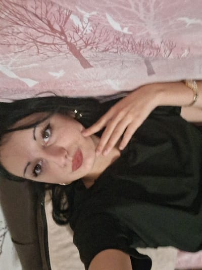 The image captures a female subject in an indoor residential setting, situated in Lanchkhuti, Georgia. The individual, identified as an adult female, possesses dark hair and light-colored eyes. Her facial features are enhanced with black eyeliner and red lipstick. She is attired in a plain black short-sleeved top. Adornments include small gold-toned hoop earrings and a gold-toned bracelet on her right wrist. The subject is depicted in a supine position, with her head oriented towards the left side of the frame, and her gaze directed towards the viewer. Her left hand is raised, with fingers resting against her chin or jawline. The background comprises a patterned textile, likely bedding or a pillow, characterized by a pink base color overlaid with a repeating motif of brown skeletal trees and stylized white bird figures. A segment of a second, solid-colored textile, light beige with a white border, is visible in the lower-left portion of the frame.