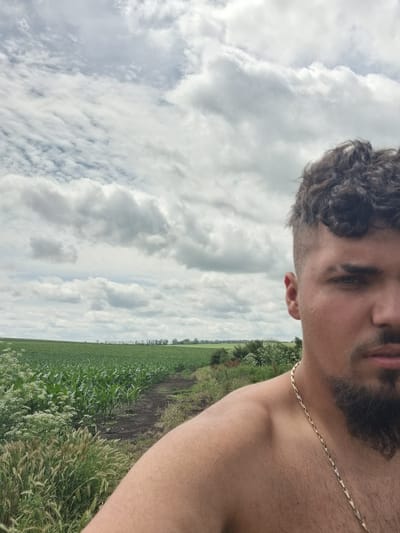 This is a close-up, selfie-style photograph taken outdoors on a cloudy day. The left two-thirds of the image shows a rural landscape with a vast, green cornfield stretching to the horizon. A dirt path runs along the edge of the field, bordered by tall grass and wildflowers. The sky is filled with dramatic, textured white and gray clouds. The right third of the image features the upper torso and right side of the face of a young man. He is bare-chested, wearing a thin, gold chain around his neck. His hair is dark and curly, styled with an undercut. He has a goatee and mustache. The photo appears to be taken in Lomnitsa, Bulgaria, a location suggested by the presence of cultivated fields common in rural Bulgarian landscapes.
