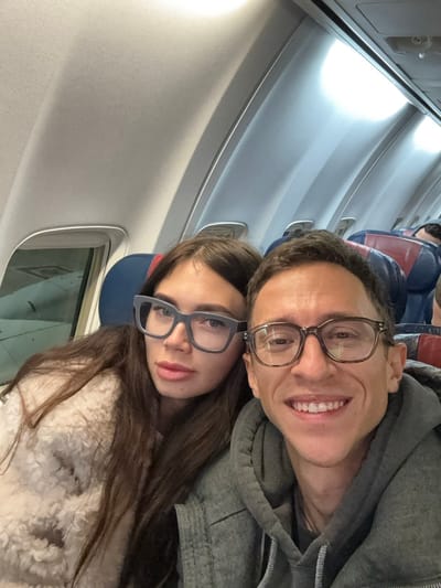 Inside an airplane cabin, a selfie captures a smiling man with glasses in a gray hoodie alongside a woman also wearing glasses. She is adorned in a light-colored, textured sweater. A window and the interior of the plane are visible. The cabin is illuminated by overhead lighting, accentuating the contours of the aircraft's walls and ceiling. The image gives the impression that the couple is seated and taking a photo during air travel.
