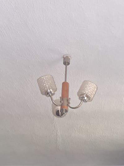 A light fixture hangs from a textured white ceiling in an indoor setting, possibly a room in Kamenka, Moldova. The fixture has a central, light brown cylindrical piece and two clear glass lampshades with a textured pattern. The lampshades are positioned on curved metal arms extending from the central body. The ceiling exhibits a rough, uneven surface with visible brush strokes or imperfections. There are no people or animals present in the image. The lighting appears to be ambient, likely from the fixture itself or other unseen sources.