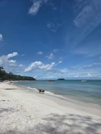 A wide, sandy beach stretches from the left foreground to the mid-right of the image, meeting the gentle, calm, turquoise ocean. The sky above is a vivid blue, dotted with scattered white cumulus clouds. In the distance, a thin, tree-covered island is visible, with a larger landmass and buildings on the far horizon. Three boats are present: a small, dark boat rests partially on the sand in the mid-left, a larger red and white boat is anchored in the mid-distance, and a white speedboat is cruising in the mid-right. Along the treeline on the left, several beach chairs are set up. In the foreground, a dark, wooden cart or barrow is partially submerged in the shallow water. The overall scene depicts a serene tropical beach on a sunny day. The location is Koh Toch Village, Cambodia.