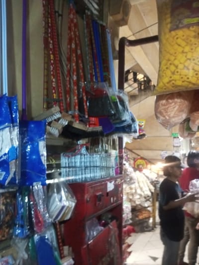A bustling market scene features a variety of goods displayed for sale. Brooms and dustpans hang vertically on the left, while bags of yellow and tan items dangle from above on the right. A red cabinet showcases stacked metallic objects. In the background, blurred figures of shoppers are visible, adding depth to the scene. The overall impression is of a busy, vibrant commercial environment.