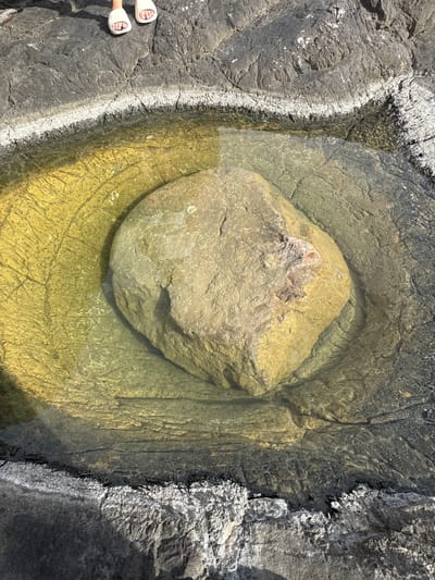 The image shows a rock formation in Seoca, Montenegro. A central, light brown rock is submerged in a shallow pool of water. The pool is surrounded by darker grey rocks. In the bottom left corner, a pair of feet wearing white sandals are partially visible. The surrounding rocks show textures of erosion and weathering.
