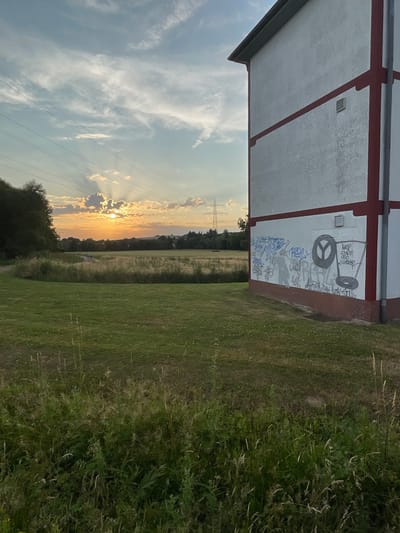 The image captures a picturesque sunset over a rural landscape in Rückershausen, Germany. The scene is dominated by a strip of mown grass on the right and a field of uncut grass and wildflowers on the left, suggesting a natural, uncultivated area. In the foreground, the white exterior wall of a building is visible, featuring red trim and what appears to be graffiti. One piece of graffiti includes a peace symbol and a drawing of a trolley, along with various tag-like scribbles in blue and grey.

The sunset is a prominent feature, casting a warm, golden light across the sky. Rays of sunlight break through scattered clouds, creating a dramatic and serene atmosphere. Power lines stretch across the sky, adding a man-made element to the natural beauty. In the distance, a tall transmission tower is visible, hinting at the infrastructure within the rural setting. The overall mood is peaceful and tranquil, enhanced by the warm tones of the sunset and the vast open space. The time of day is clearly evening, as indicated by the setting sun. There is no clear indication of any specific activity or event taking place beyond the natural transition from day to night.