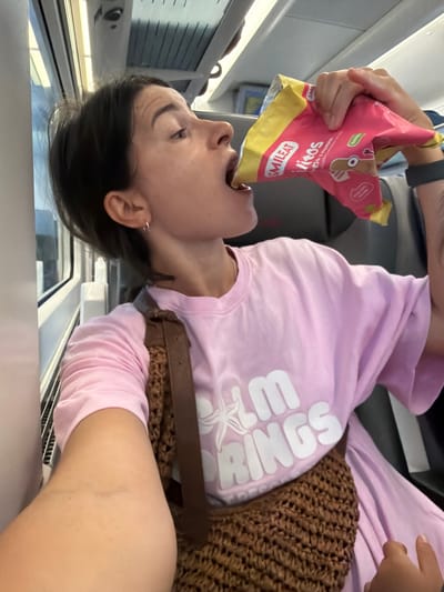 The image depicts a woman inside a train carriage, in the city of Tarragona, Spain. She is wearing a pink t-shirt with the text "PALM SPRINGS" printed in white. A woven brown bag is slung over her shoulder, resting on her chest and stomach. The woman is in the process of eating from a pink and yellow bag of snacks, holding it up to her mouth. The interior of the train carriage is visible, with seats and windows. The lighting suggests it is daytime.