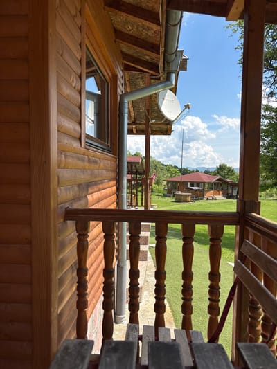The image depicts a static outdoor scene captured from the perspective of a wooden porch or balcony, likely at a cabin or lodge in Zasele, Bulgaria. The immediate foreground shows the interior of this wooden structure, with a partial view of a rustic wooden table or bench in the bottom-center. A prominent wooden balustrade and handrail, crafted with turned balusters, extends across the lower portion of the frame, defining the edge of the porch.
To the left, the exterior wall of the building is constructed from horizontal logs, showcasing a light brown wood grain. A window with dark framing is set into this wall. Above the window, a gray metal rain gutter system is visible, running horizontally and then descending vertically. Attached below the gutter, a white satellite dish, bearing the "Бусатком" (Bulsatcom) logo, is mounted and points upwards towards the sky. The overhang of the porch roof is visible at the top, constructed with exposed wooden beams and plywood sheathing.
Beyond the porch and railing, a vibrant green lawn, possibly artificial turf or meticulously maintained grass, stretches into the midground. In the center of the midground, a substantial single-story wooden building with a prominent red tile roof stands. This building features numerous windows and a covered porch area. To its left, a circular wooden structure, resembling a hot tub or a large planter, is situated on the lawn, adjacent to a wooden swing set. A tall, thin metal pole is visible behind the hot tub structure, extending upwards into the sky.
The background consists of rolling hills or mountains covered with dense green foliage, indicating a rural or semi-forested environment. The sky above is clear blue with scattered, fluffy white cumulus clouds. No people or dynamic actions are present in the frame.