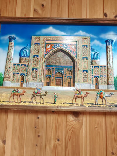 A detailed relief artwork depicts a mosque with two tall minarets and blue domes against a bright blue sky with white clouds. The mosque features intricate geometric patterns and Arabic calligraphy in shades of blue, gold, and orange. In front of the mosque, a caravan of three camels walks across a sandy terrain. Two camels are ridden by figures in traditional attire, each carrying colorful saddlebags. A third camel is being led by a standing figure holding a staff. The artwork is framed by a wooden border and mounted on a wooden wall with vertical planks.