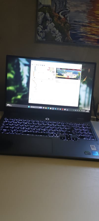 A black laptop with illuminated keys is open on a light-colored desk. The laptop screen displays a white window with a list of items and icons, and a smaller window showing a landscape image with text overlaid, suggesting a streaming or gaming interface. To the left of the screen, a portion of a digital wallpaper with trees is visible. In the upper background, a painting hangs on a wall, depicting a stone structure by a body of water with warm-toned reflections. A power cord is visible plugged into the side of the laptop. The scene appears to be indoors, with the wall having a textured finish. No individuals are present in the image. The image does not provide any information regarding the city of Skopin, Russia.